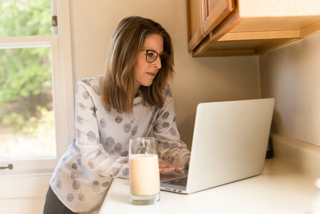 Older woman with glasses and long hair on a laptop working, glass of milk next to her