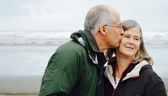 Older man in a hunter green jacket kissing a woman on the cheek with beach and waves behind them