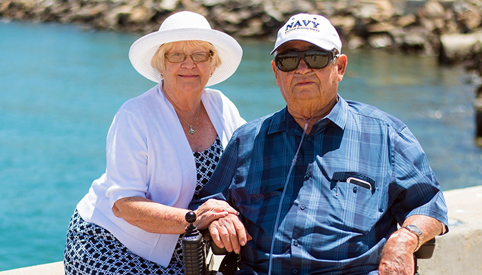 Elderly couple sitting by the water, woman in white blouse and hat, man in wheelchair and Navy baseball hat and sunglasses